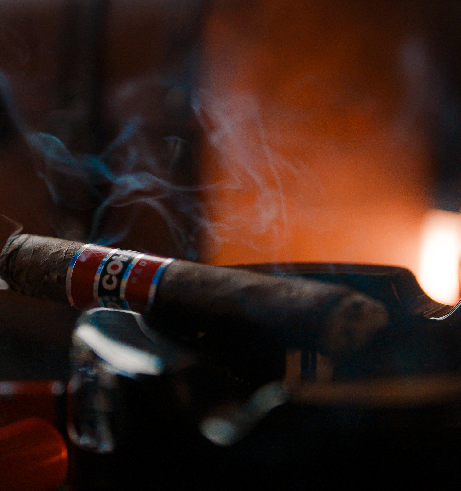 A cigar rests in an ashtray, with smoke rising. A glass is nearby, creating a warm atmosphere.