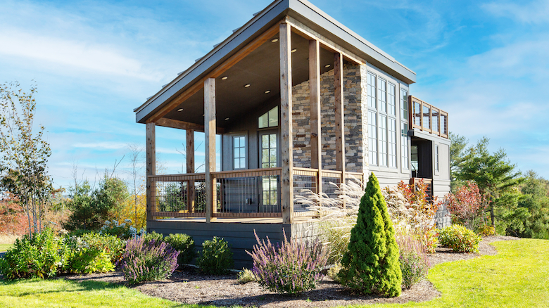 A modern tiny house with large windows and a wooden porch, surrounded by a neatly landscaped garden under a clear blue sky.