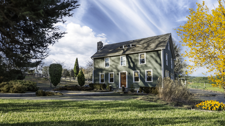 A green two-story house surrounded by trees and shrubs under a blue sky with wispy clouds, with a mowed lawn and colorful foliage.