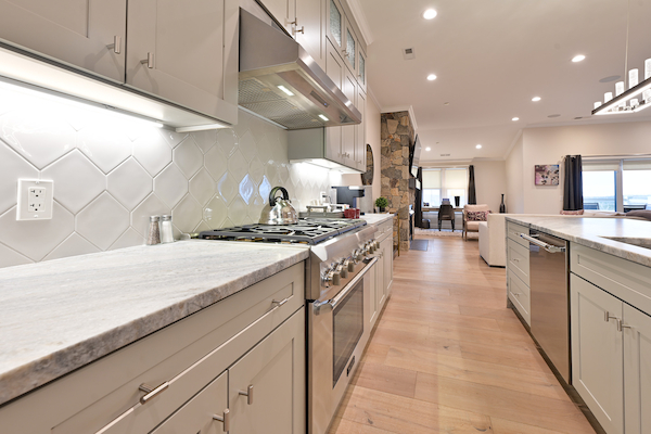 A modern kitchen with light-colored cabinets, marble countertops, stainless steel appliances, and a dining area visible in the background.