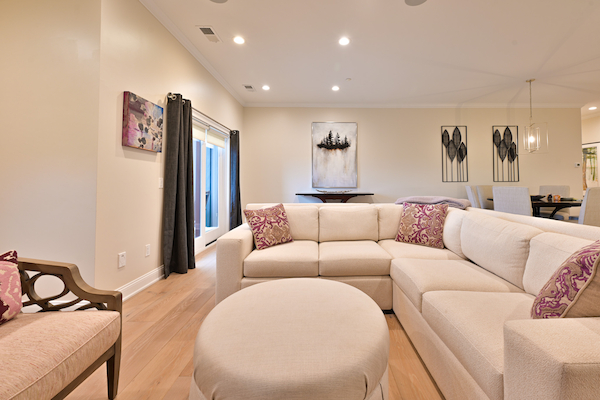 A modern living room with beige sofas, a matching ottoman, contemporary artwork, wooden flooring, and ceiling lights in an open space layout.