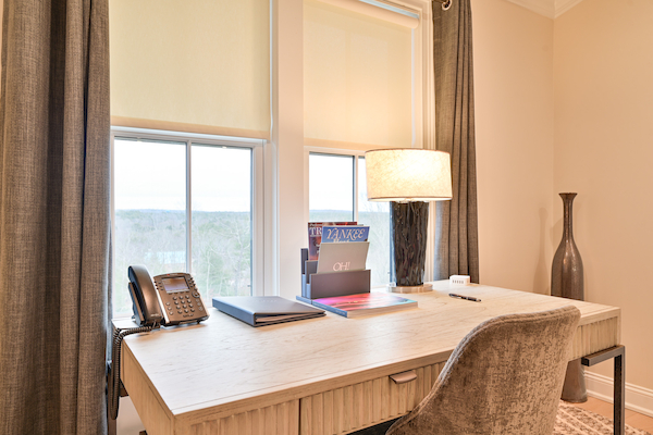 A modern home office features a desk with a phone, notebook, magazines, lamp, and decorative vase near a window with beige curtains.