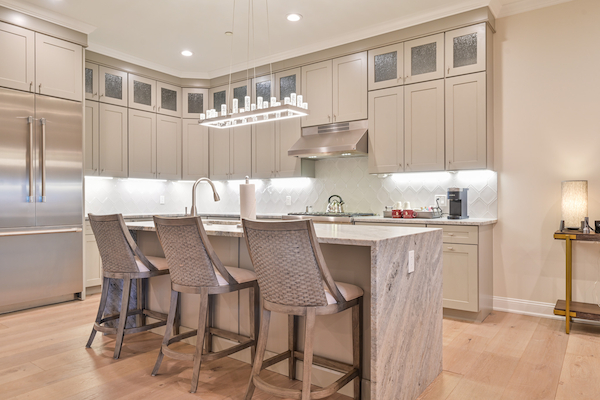 A modern kitchen featuring light-colored cabinets, a large island with four bar stools, stainless steel appliances, and a sleek overhead light fixture.