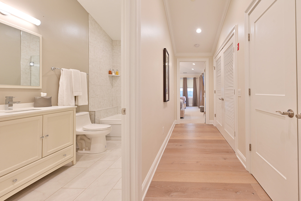 The image shows a hallway leading to a room, with a bathroom on the left. The flooring is light wood, and the bathroom has a modern design.