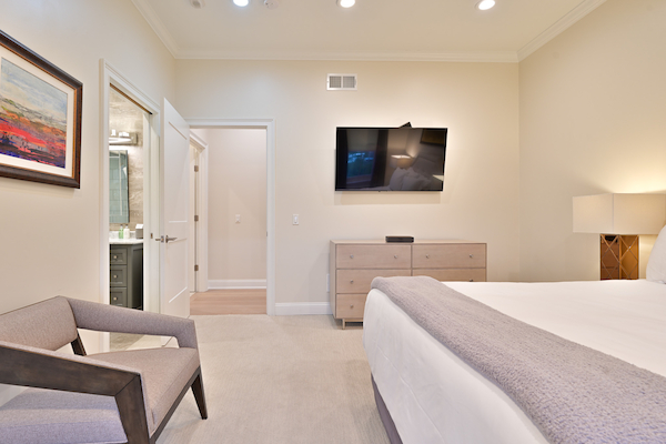 A modern bedroom with a wall-mounted TV, dresser, chair, lamp, and artwork. There’s a bed with white bedding and a doorway leading to a bathroom.