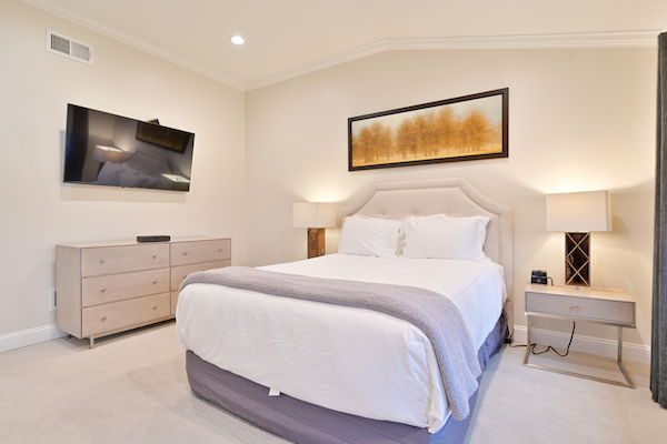 A neatly arranged bedroom with a bed, dresser, wall-mounted TV, two bedside tables with lamps, and a framed artwork above the bed.