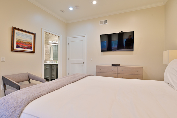 The image depicts a modern bedroom with beige walls, a wall-mounted TV, a dresser, a chair, a bed with white linens, and an adjoining bathroom.