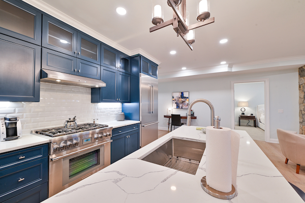 A modern kitchen with blue cabinets, a stainless steel stove, marble countertop, and sink; adjacent to a dining area and a view of another room.