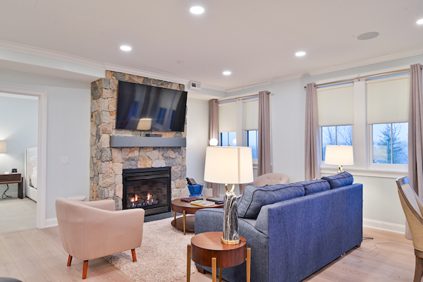 A modern living room with a stone fireplace, wall-mounted TV, blue sofa, beige armchair, and side tables with lamps, adjacent to a bedroom.