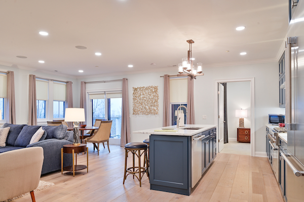 The image shows a modern, open-concept living space with a kitchen, dining area, and seating area. The design features light wood floors.