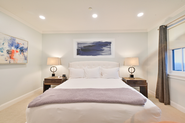 A neatly arranged bedroom with a bed, two bedside tables with lamps, abstract art on the walls, and a large window with curtains.