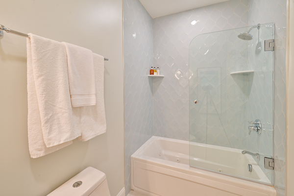 A bathroom with a glass-enclosed bathtub, two white towels on a rack, and a small shelf holding toiletries above the bathtub.