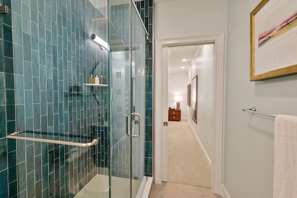 A bathroom with a glass-enclosed shower, teal tiles, a towel rack, and a view into a hallway with furnishings and artwork.