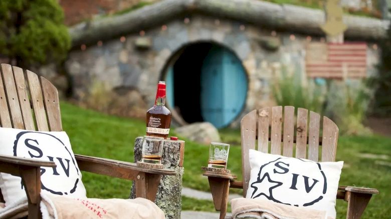 Two wooden chairs with pillows and a small table holding a bottle and two glasses in front of a Hobbit-like house with a blue door.