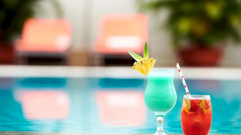 Two colorful cocktails with garnishes sit poolside, with lounge chairs and greenery in the blurry background.