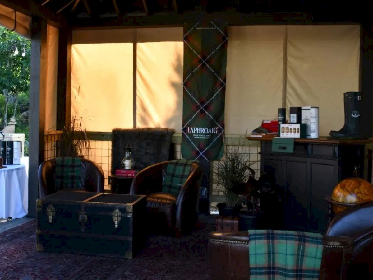 The image shows a cozy lounge setup with leather chairs, a trunk as a table, a globe, and a shelf with various items. Laphroaig signage is visible.