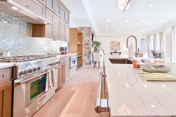 A modern kitchen with a large island, gas stove, wooden cabinetry, tiled backsplash, and a dining area in the background.