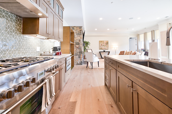 A modern kitchen with stainless steel appliances, wooden cabinets, a large island, and a stylish hexagon backsplash; an open living area beyond.