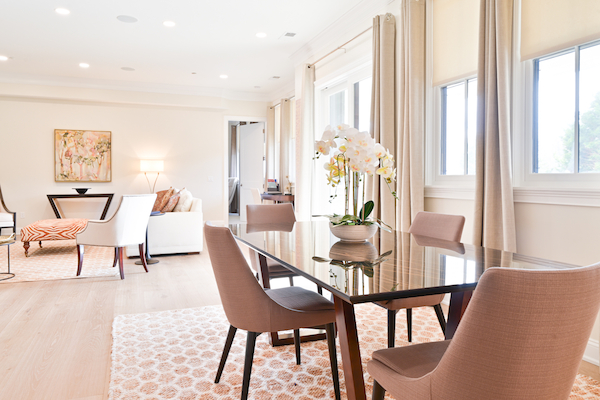 A modern dining area with a glass table, pink chairs, and a potted orchid, adjoining a living space with a sofa, chairs, and wall art.