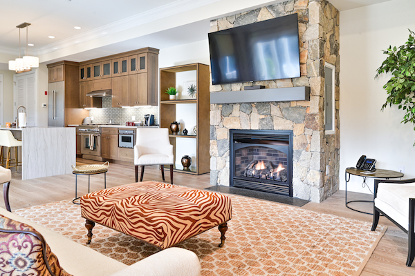 A modern living room with a stone fireplace, flat-screen TV, patterned ottoman, and open kitchen in the background ends the sentence.