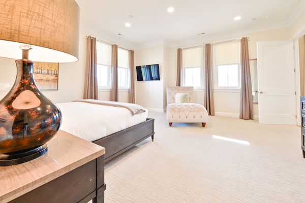 A bright bedroom with a bed, lamp, windows, TV, and a chaise lounge chair in a mostly white and beige color scheme and carpeted floor, ending the sentence.