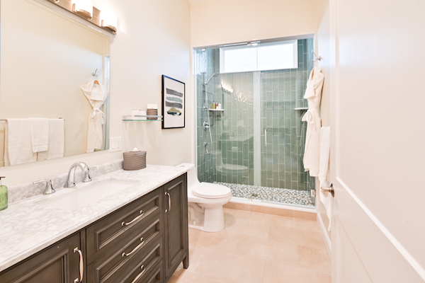 This image shows a modern bathroom with a vanity, sink, toilet, and a glass-enclosed shower with green tiles and pebble flooring.