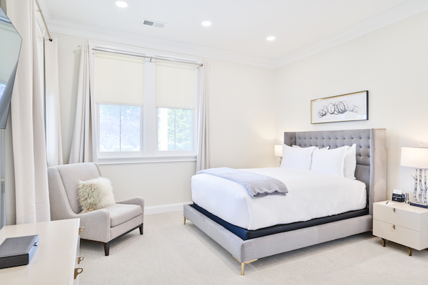 A bright bedroom with a bed, armchair, nightstand, lamps, and a wall picture. There's also a window with blinds and curtains.