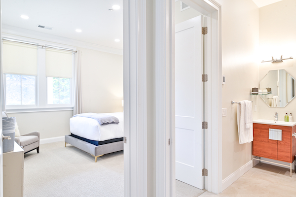 The image shows a bedroom with a bed, chair, and window, adjacent to a bathroom featuring a sink, mirror, and towel.