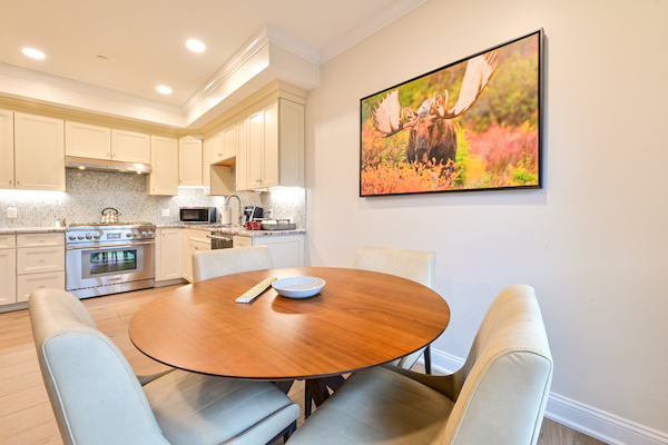 A modern kitchen and dining area features a round wooden table with four chairs, and a wall-mounted photo of a moose in nature.