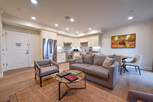 A modern living room with a gray sofa, armchair, coffee table, kitchen area, dining set, and a landscape painting on the wall.