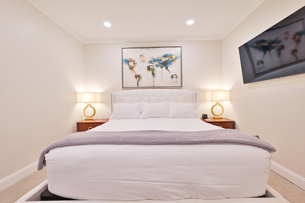 A neatly arranged bedroom with a white bedspread, side tables with lamps, a large wall art above the bed, and a TV mounted on the wall.