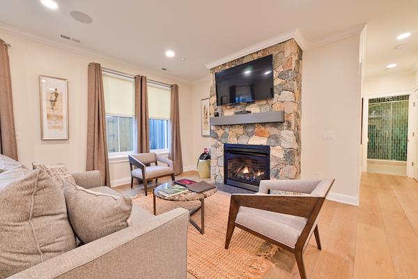 A cozy living room features a stone fireplace, wall-mounted TV, light-colored sofas, and wooden chairs, with a hallway visible in the background.