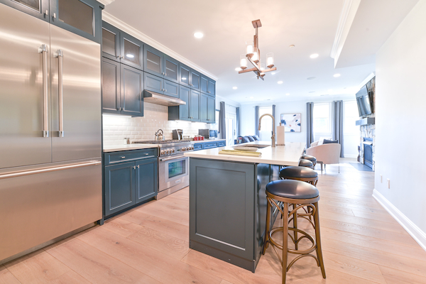 A modern kitchen with stainless steel appliances, blue cabinets, an island with stools, and hardwood flooring, opening to a bright living area.