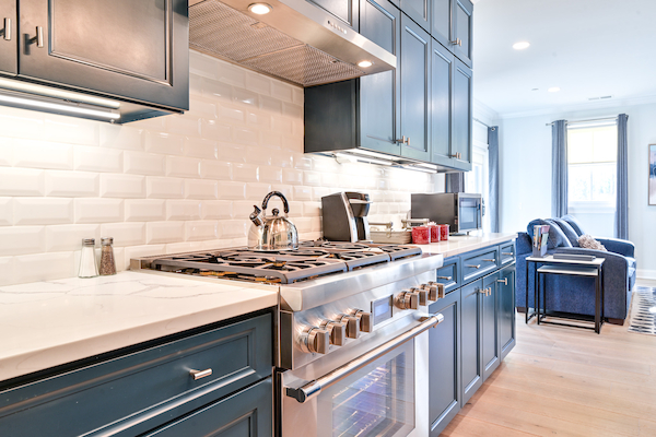 A modern kitchen with blue cabinets, white backsplash, stainless steel stove, kettle, and cooking utensils. A living room is visible in the background.