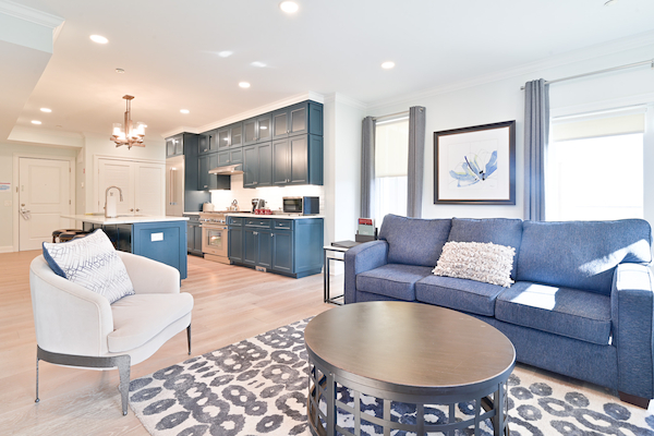 A modern living room and kitchen area with a blue sofa, gray armchair, round coffee table, patterned rug, and dark blue cabinets in the kitchen.