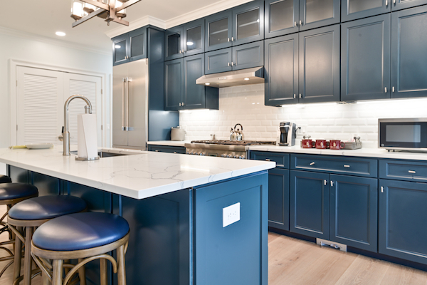 A modern kitchen with blue cabinets, a white island, bar stools, stainless steel appliances, and under-cabinet lighting.