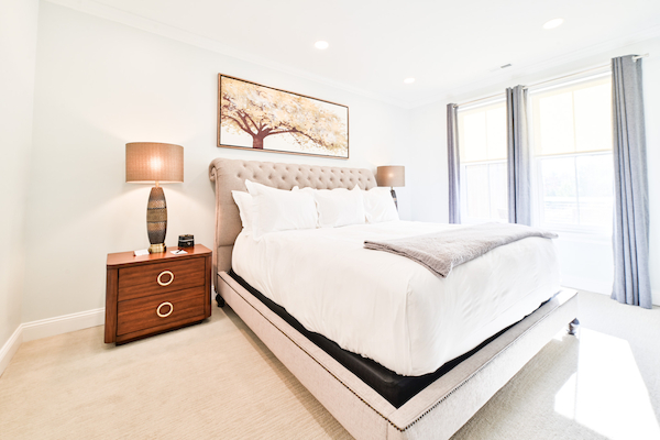 A bright, modern bedroom with a large bed, two nightstands with lamps, and a tree painting above the bed. The room has natural light.