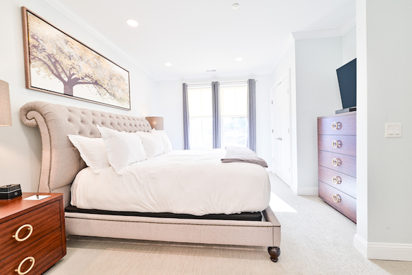 A modern bedroom with a large bed, a wooden nightstand, a dresser with a TV, and a tree painting above the bed. Bright natural light through windows.