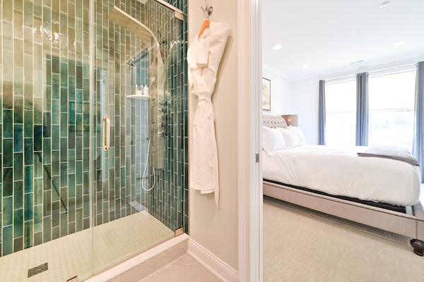 The image shows a view of a bathroom with a glass-door shower and a bedroom with a neatly made bed and large windows with curtains.