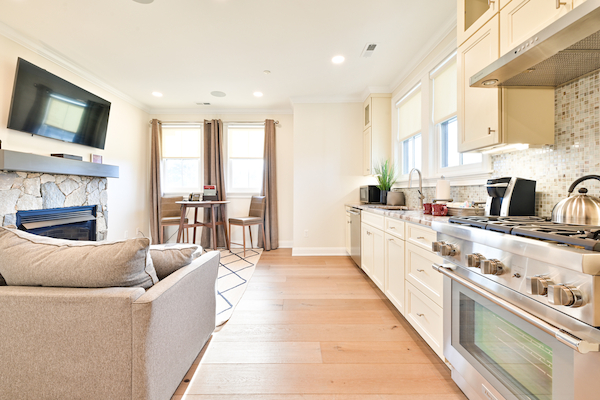 A modern kitchen and living area with light-colored cabinetry, a grey sofa, a dining table, a flat-screen TV, and hardwood floors.