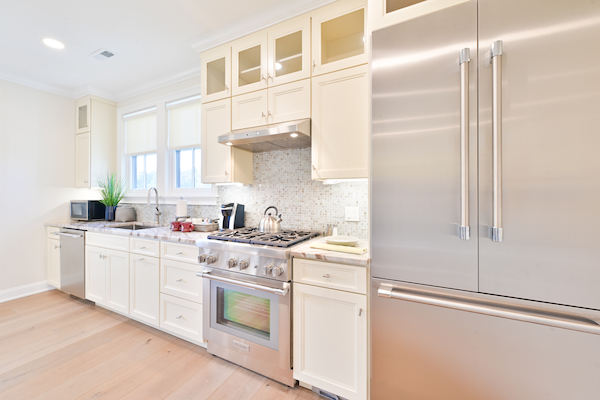 This image shows a modern kitchen with white cabinetry, a stainless steel refrigerator, an oven with a stove, a sink, and various appliances.