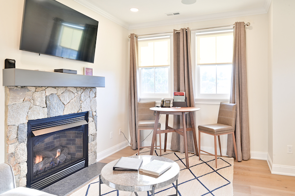 The image shows a cozy living room with a stone fireplace, a wall-mounted TV, a small round table, two chairs, and a window with curtains.