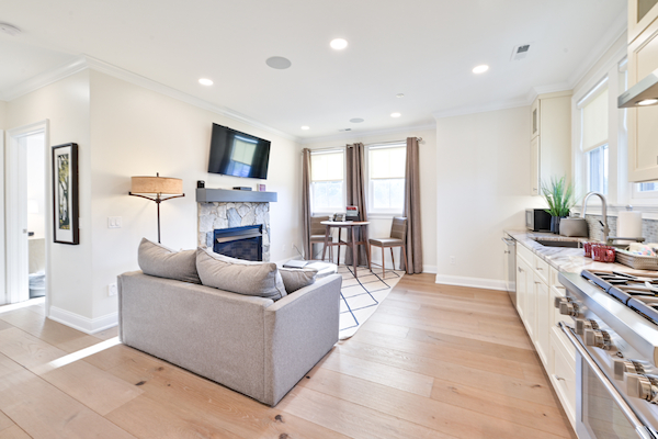 A modern living room with a wall-mounted TV, fireplace, gray couch, kitchen area, and dining table with chairs, all in a bright, well-lit space.