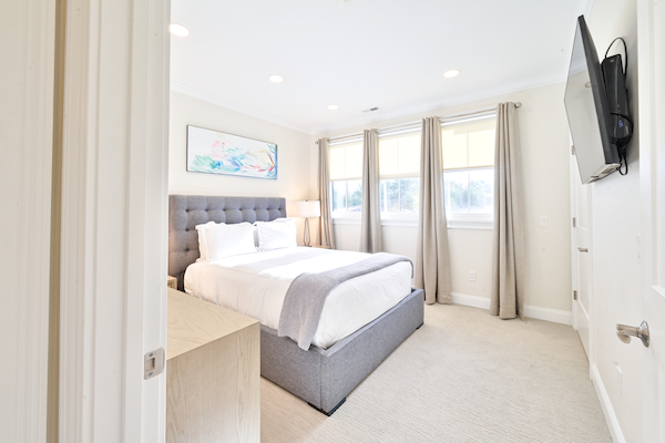 A bright bedroom with a neatly made bed, a wall-mounted TV, artwork above the bed, and large windows with curtains.