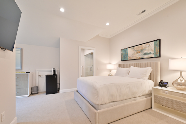 A modern bedroom with a white bed, two nightstands with lamps, a wall-mounted TV, dresser, mini-fridge, and a bathroom in the background.