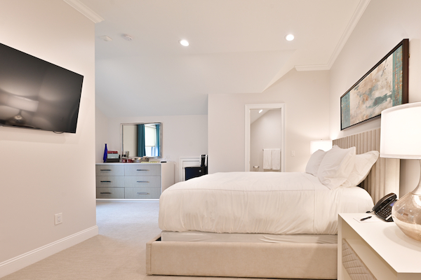 A bright, modern bedroom with a large bed, wall-mounted TV, dresser, abstract art, and a nightstand. There's an open doorway leading to a bathroom.