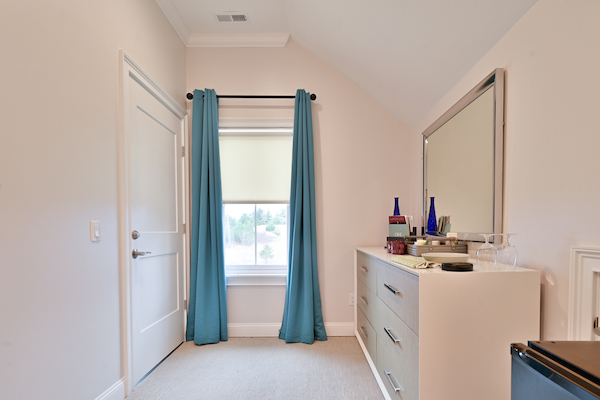 A room with a window and blue curtains, a door, a dresser with a mirror above, and various items on top of the dresser, ending the sentence.