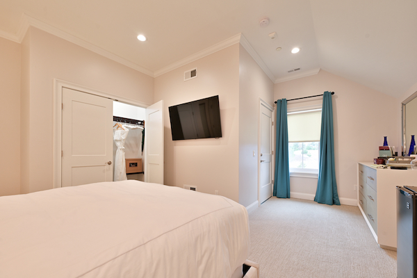 The image shows a bedroom with a bed, wall-mounted TV, dresser, closet, white walls, ceiling lights, blue curtains, and a window ending the sentence.