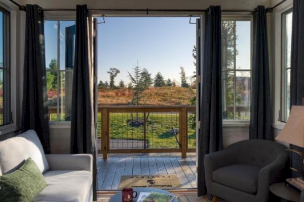 A cozy living room with a gray couch, armchair, and a lamp, opening to an outside deck with a scenic view of trees and greenery.