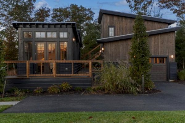 A modern, two-story wooden house with large windows, a front porch, a tree, and a paved driveway, against a backdrop of trees.